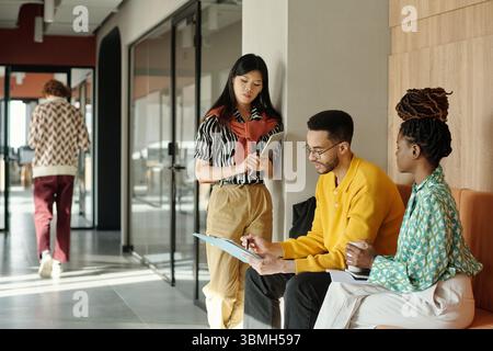 Young adult Asian businesswoman standing with notebook talking to Black colleagues sitting on bench reviewing documents in modern coworking office hallway Stock Photo