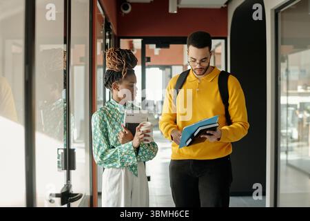 Young adult Black woman holding coffee cup and documents standing next to man with glasses wearing yellow sweater and backpack using digital tablet Stock Photo