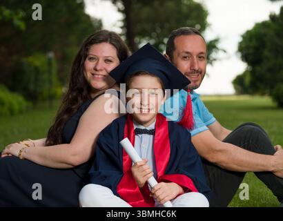 Primary school graduation A boy in his graduation outfit sits on the grass with his parents. The family looks happily at the camera. Stock Photo