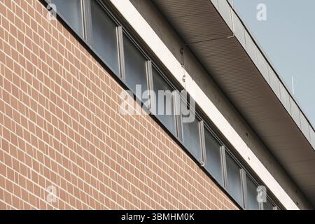 Red brick wall rising to meet metal roof and windows on a modern building, creating strong lines and contrast Stock Photo