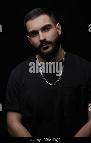 A close-up dramatic portrait of a young serious guy, musician, singer, rapper with a beard and arms crossed on his chest on a black isolated backgroun Stock Photo