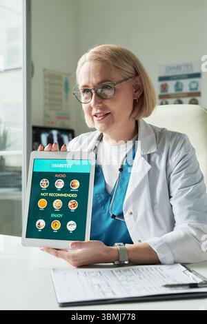 Middle aged Caucasian female doctor showing digital tablet with diabetes dietary recommendations to patient during medical consultation Stock Photo
