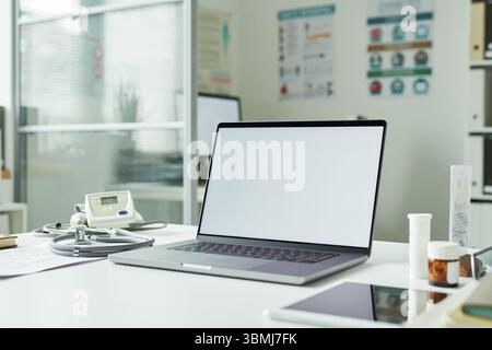 Medical office desk displaying open laptop with blank screen, digital blood pressure monitor, stethoscope, prescription medication bottles, diabetes p Stock Photo