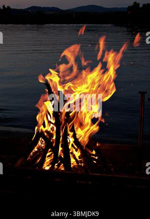Campfire glowing at night on the riverside, with mountain silhouettes in the background Stock Photo