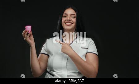 Woman smiling while holding and pointing at a pink menstrual cup against an isolated black background, promoting eco-friendly feminine care solutions Stock Photo