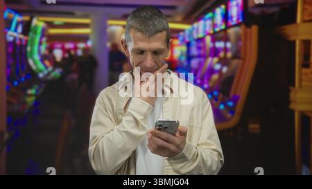Caucasian man using smartphone in a vibrant casino, surrounded by colorful slot machines, focusing on his device indoors. Stock Photo