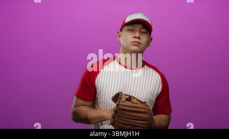 Young chinese man in baseball uniform with glove stands against isolated pink background with confident expression. Stock Photo
