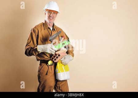 Smiling construction worker dressed in work clothes holding tools and building materials isolated on brown background. Front view. Stock Photo