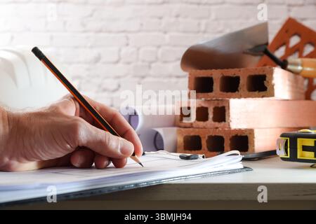 Detail of architect writing down in a folder with documents on white wooden table with tools and construction materials. Front view. Stock Photo