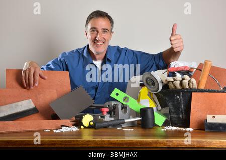 Smiling tiler with ok gesture showing tools and materials on wooden table. Front view. Stock Photo