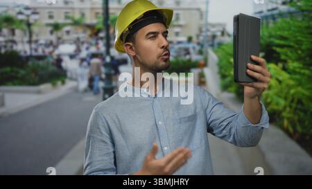 Young man in hardhat making video call on city street using tablet showcasing outdoor urban environment and construction concept. Stock Photo