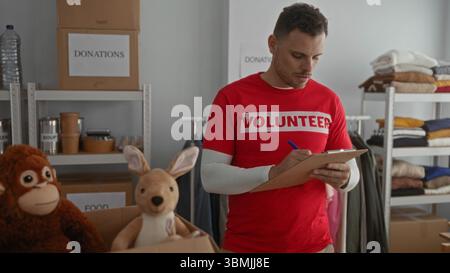 Young man wearing red volunteer shirt checking items on clipboard in charity donation room filled with clothes and toys. Stock Photo