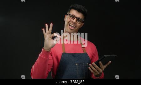 Young man with glasses and apron interacts with tablet against dark background showcasing modern technology and casual fashion style mood highlighting Stock Photo