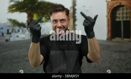 Young man with beard gestures humorously with middle fingers on a city street while wearing black gloves and a striped apron, appearing amused in an o Stock Photo