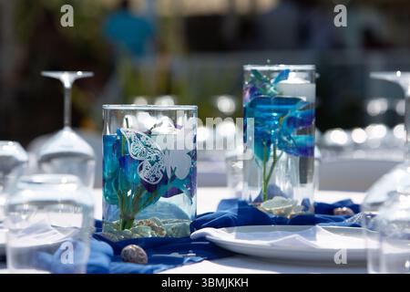 Wedding reception table is decorated with blue flowers, butterfly decorations, seashells, and elegant glassware in outdoor setting Stock Photo