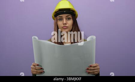 Young woman architect wearing yellow hardhat examines blueprint against purple background with focus and concentration, showcasing engineering skills Stock Photo