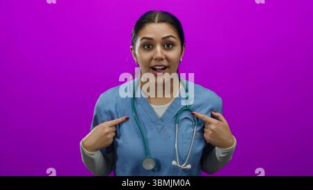 Hispanic nurse in blue scrubs with a stethoscope looks surprised, pointing at herself against an isolated pink wall background. Stock Photo
