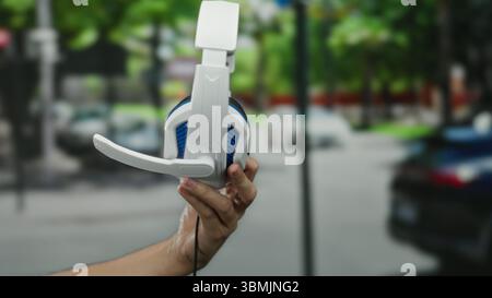 Man holding white headphones with microphone in urban city street background showcasing technology outdoors. Stock Photo