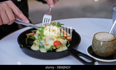 Person enjoying eggs benedict on a table with cappuccino in a sunny cafeteria showcasing vibrant salad and creamy sauce. Stock Photo