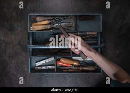 Hand holding vintage cold chisel above old blue metal toolbox filled with assorted tools from above Stock Photo