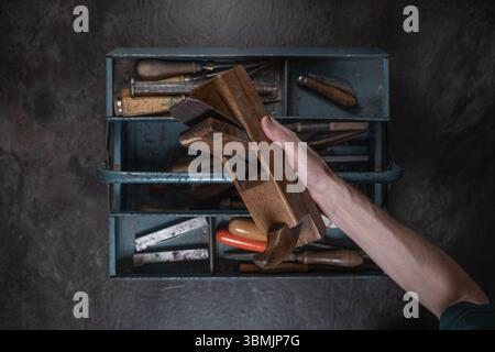 Top-down view of hand holding traditional wooden hand plane above open toolbox with various tools on rustic dark surface. Stock Photo