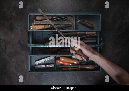 Top-down view of a hand gripping screwdriver above open toolbox filled with various tools on rustic dark surface. Stock Photo