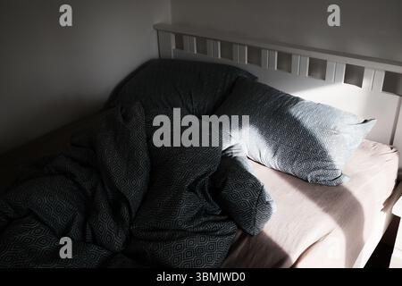 Moody bedroom scene with sunlight casting shadows on a crumpled patterned bed. The morning sunlight from the window falls on the unmade bed Stock Photo