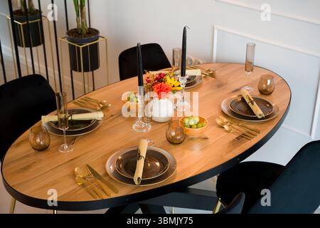 Elegantly set wooden oval dining table in a modern dining room. The table is prepared for a festive dinner party with dark plates, gold cutlery, and s Stock Photo