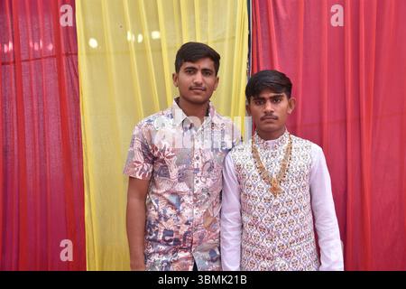 Two Young Indian Men Posing Together at Wedding Function with Colorful Drapes in Background Stock Photo
