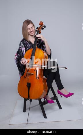 Adult Caucasian woman in studio playing cello, sitting, smiling, wearing floral kimono, black pants, pink heels. Stock Photo