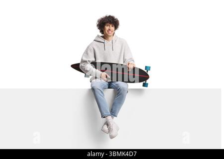 Guy holding a longboard and sitting on a blank panel isolated on white background Stock Photo