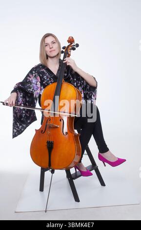 Caucasian woman in floral kimono robe plays cello in a studio setting. Sitting pose, wearing black pants and pink heels. Stock Photo