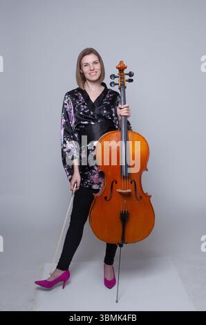Caucasian woman, 30s, standing in studio with cello & bow. Smiling, wearing floral kimono top, black pants, pink heels. Full length shot. Stock Photo