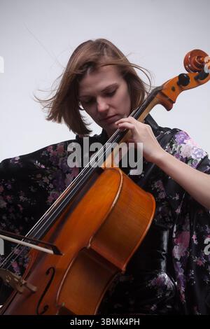 Young adult Caucasian woman playing cello in studio. Wearing black floral kimono, focused on instrument. Stock Photo