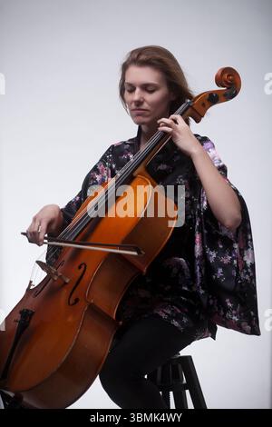 Adult Caucasian woman playing cello in studio. Sitting, wearing black floral kimono robe, eyes closed, focused. Stock Photo