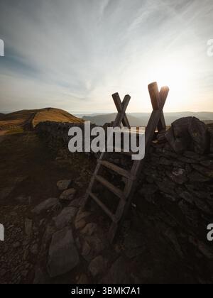Helvellyn, Wooden ladder stile over dry stone wall in Lake District landscape at sunset, traditional countryside footpath crossing. Stock Photo