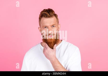 Handsome young man with a red beard poses thoughtfully in a casual white t-shirt against a pink background Stock Photo