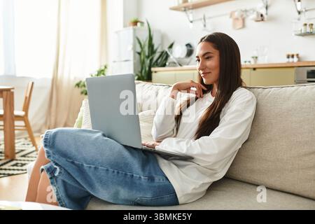 Beautiful woman relaxes on the couch, engaged with her laptop in a cozy home environment. Stock Photo