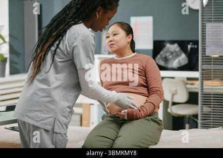 Asian pregnant young adult woman sitting on examination table interacting with Black woman healthcare professional during prenatal checkup in medical clinic setting Stock Photo