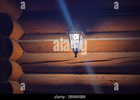 Street lamp hanging on the wall brightly illuminates the red painted logs of the tree in the late evening. Stock Photo