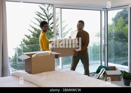 Diverse couple carrying large cardboard moving box in modern bedroom, with bubble wrap Stock Photo