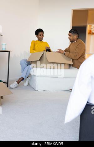 Unpacking couple sitting on white mattress in mostly empty room, handling items from cardboard box Stock Photo