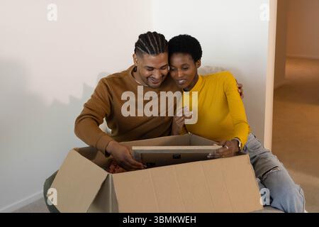 Unpacking diverse couple leaning on carpeted floor in empty room, with cardboard box and artwork Stock Photo