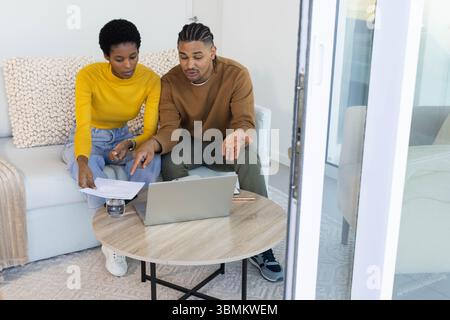 Reviewing diverse couple sitting on sofa in bright living room, with laptop documents pencil Stock Photo