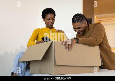 Unpacking Diverse couple opening large box on white surface in simple room, with smartphone Stock Photo
