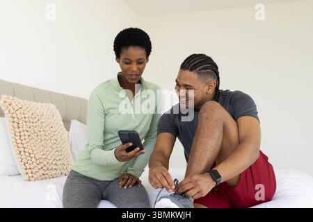 Sitting diverse couple using smartphone while tying running shoes on bed in bedroom, with pillow Stock Photo