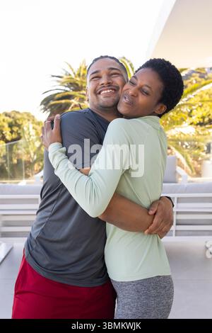 Hugging diverse couple in sportswear embracing on balcony patio, with glass railing and palm trees Stock Photo
