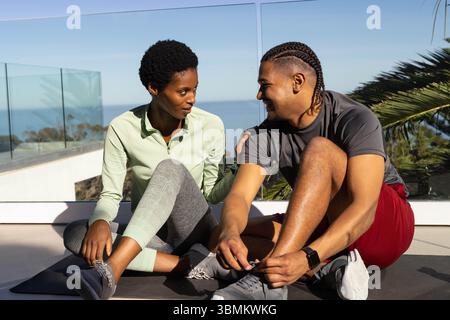 Practicing yoga, diverse couple in sportswear on seaside terrace, with exercise mats and ocean view Stock Photo