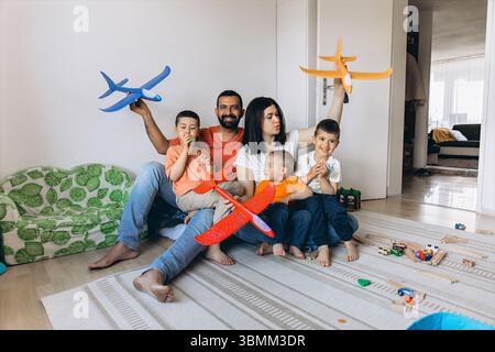 A happy family of five sits barefoot on a rug in a bright room, playing with toy airplanes and wooden trains, enjoying joyful, relaxed moments of toge Stock Photo