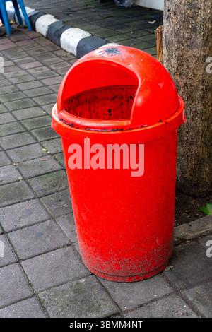 Red trash can is sitting on the sidewalk. The trash can is empty and has a black lid Stock Photo